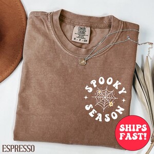May include: Espresso-coloured t-shirt with the words "Spooky Season" in a circular design, featuring a spiderweb and flowers. A silver necklace with a pendant rests on the shirt. The shirt is folded and placed on a white surface.