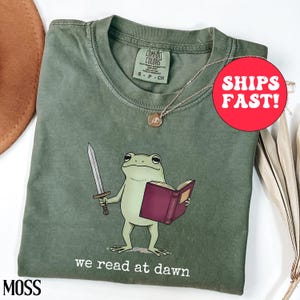 May include: Moss green t-shirt with a cartoon frog holding a sword and a book. The frog is reading the book with the text "we read at dawn" below. A gold necklace with a pendant is on the shirt.