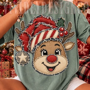 Puede incluir: Una camiseta verde salvia con una gran aplicación de estilo desgastado de un reno con un gorro de Papá Noel a rayas rojas y blancas. El reno tiene pelaje marrón, nariz roja, astas y un detalle de estrella en el pompón.