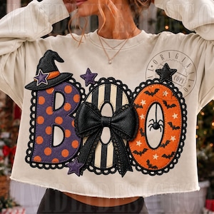 May include: A cream-colored long-sleeve sweatshirt featuring a large graphic that spells BOO. The B is purple with orange polka dots and a witch hat, the O is black and white striped with a black bow, and the O is orange with bats and stars.