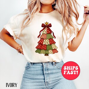 Op de afbeelding: Ivoor kleurig T-shirt met een patchwork kerstboom ontwerp. De boom is versierd met rood, groen en wit patroonstof, een rode strik en een gouden ster. De tekst "IVORY" staat onderaan het shirt.