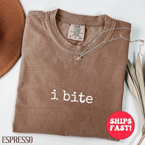 May include: Espresso-colored t-shirt with the text "i bite" in white. The shirt has a crew neck and short sleeves. A silver necklace with a small pendant is draped across the shirt. A brown hat is visible in the upper left corner.