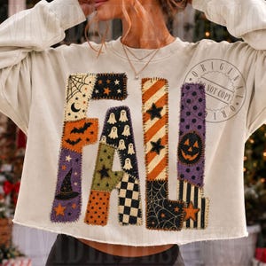 May include: A cream-coloured long-sleeve sweatshirt featuring the word FALL in large, block-style letters. Each letter is composed of various fabric patches with Halloween-themed patterns including ghosts, bats, pumpkins, stars, and spiderwebs.