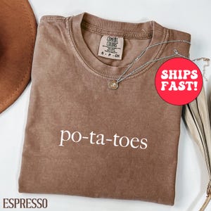 Könnte beinhalten: Espressofarbenes T-Shirt mit dem Wort "po-ta-toes" in Weiß. Das Shirt hat einen Rundhalsausschnitt und ein kleines Etikett am Ausschnitt. Eine silberne Halskette mit Anhänger liegt über dem Shirt. Ein roter Kreis mit der Aufschrift "SHIPS FAST!" ist sichtbar.