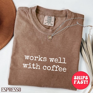 Puede incluir: Camiseta color espresso con la frase "works well with coffee" en letras blancas. La camiseta tiene cuello redondo y mangas cortas. Un collar plateado con un pequeño colgante descansa sobre la camiseta. Un sombrero marrón es parcialmente visible.