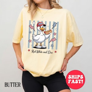 May include: A pale yellow t-shirt with a cartoon duck wearing a red cap and holding a hot dog. The graphic includes stars, lightning bolts, and the text "Red White and Due". The word "America" is on the cap.
