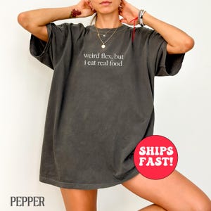 Puede incluir: Una camiseta gris oscura de gran tamaño con el texto "weird flex, but i eat real food" impreso en blanco en la parte delantera.