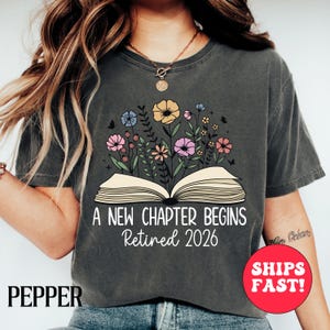 Könnte beinhalten: Dunkelgraues T-Shirt mit einem Blumendesign, das aus einem offenen Buch hervorgeht. Der Text lautet "A NEW CHAPTER BEGINS Retired 2026". Die Blumen sind in Rosa, Gelb und Blau gehalten. Das Shirt hat eine lockere Passform.