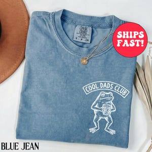 Puede incluir: Camiseta Comfort Colors azul vaquero con un gráfico blanco de una rana sosteniendo una bebida y el texto "COOL DADS CLUB". La camiseta tiene cuello redondo y mangas cortas. También se ve un collar dorado con un colgante.