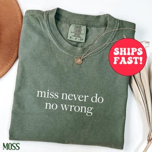Op de afbeelding: Een mosgroen t-shirt met de witte tekst "miss never do no wrong". Een goudkleurige ketting met hanger ligt over het gevouwen shirt. Het shirt heeft een label van Comfort Colors.