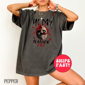 May include: Dark gray t-shirt with the text "IN MY SLASHER ERA" in red. The graphic features a half-face mask with blood splatter and a few small bats. The shirt is a relaxed fit.