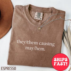 May include: Espresso-colored t-shirt with the text "they/them causing may/hem" in white. The shirt has a crew neck and short sleeves. A silver necklace with a pendant is draped across the shirt.
