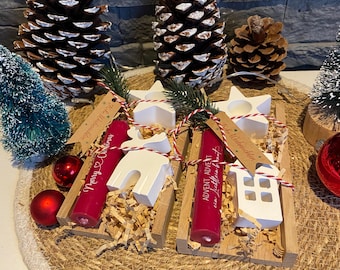 May include: Christmas-themed arrangement featuring two wooden trays with red candles, white star and house-shaped candle holders, and decorative accents. The candles have text that reads "Merry Christmas" and "Advent". The arrangement includes pine cones and ornaments.