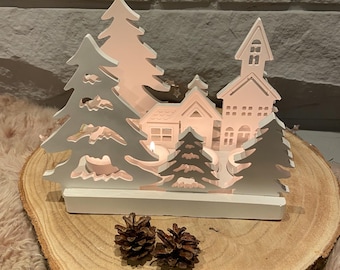 Puede incluir: Portavelas de madera blanca en forma de pueblo navideño con una iglesia, casas y árboles. El portavelas tiene recortes para velas de té y está colocado sobre una rodaja de madera.