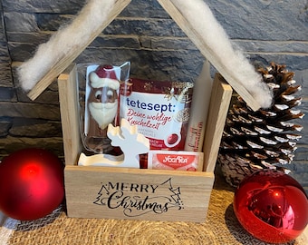 May include: A wooden house-shaped gift box with a "Merry Christmas" inscription. Inside are a chocolate Santa, tea, and other treats. Red ornaments and a pine cone decorate the scene, creating a festive holiday display.