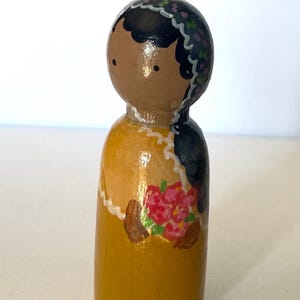 Puede incluir: Una muñeca de madera pintada a mano con piel marrón, cabello negro y un vestido amarillo. La muñeca lleva una diadema floral y tiene un ramo de flores rosas pintadas en su vestido.