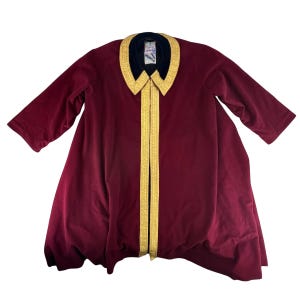 May include: A burgundy velvet robe with a black collar and gold trim. The robe has three-quarter sleeves and a flowing design. The gold trim runs down the front. A tag is visible on the collar.