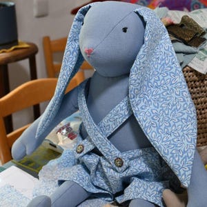 Könnte beinhalten: Ein blaues Stoff-Hasenspielzeug, das einen Overall mit einem weißen und blauen Blumenmuster trägt. Der Hase hat lange Ohren und eine rosa Nase.
