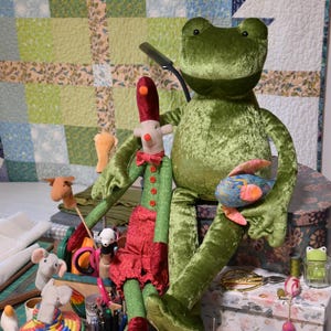 May include: A large green velvet frog doll and an elf doll are displayed with craft items. The elf has a red hat, green jacket, and red skirt. Other items include a sewing box, pincushions, and a colourful basket.