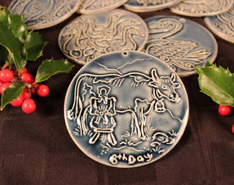 May include: A blue ceramic ornament featuring a white cow and the text "8th Day" against a dark background.