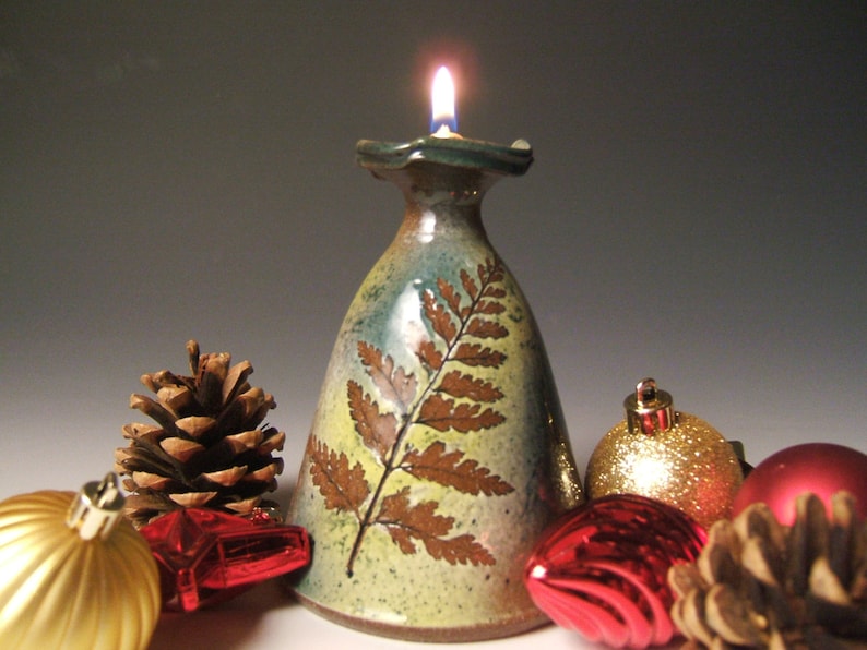 May include: A ceramic oil lamp with a fern design in shades of brown, green, and yellow. The lamp is lit and surrounded by pine cones and Christmas decorations.