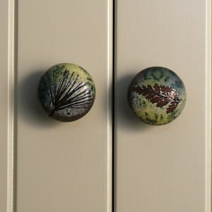 May include: Two ceramic cabinet knobs with a green and brown glaze. One knob features a pine tree design, while the other has a fern leaf design.