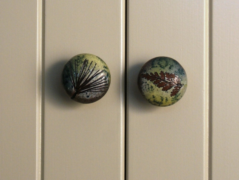 May include: Two ceramic cabinet knobs with a green and brown glaze. One knob features a pine tree design, while the other has a fern leaf design.