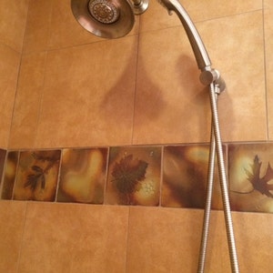 Könnte beinhalten: Ein Duschkopf mit silberner Oberfläche und einem flexiblen Schlauch ist an einer gekachelten Wand montiert. Die Fliesen sind braun mit einem Muster aus Blättern in Braun-, Grün- und Gelbtönen.