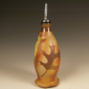 May include: A ceramic olive oil bottle with a metal pour spout. The bottle is decorated with a brown leaf design on a tan background.