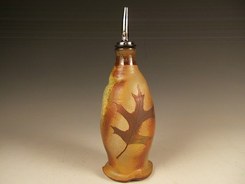 May include: A ceramic olive oil bottle with a metal pour spout. The bottle is decorated with a brown leaf design on a tan background.