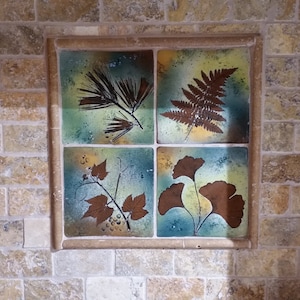 Puede incluir: Cuatro azulejos cuadrados de cerámica con un diseño decorativo de hojas prensadas en tonos marrones, verdes y azules. Los azulejos están enmarcados con piedra beige.