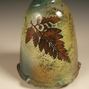 May include: A ceramic vase with a green, brown and yellow glaze. The vase features a fern design in brown on the front.