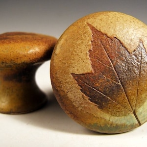 May include: Two ceramic cabinet knobs with a rustic brown glaze. One knob features a pressed leaf design in shades of brown and red.