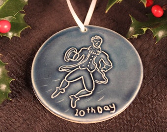 May include: A blue ceramic Christmas bauble featuring a white silhouette of a man in a hat and coat, holding a hat in his hand. The bauble reads "10th day".