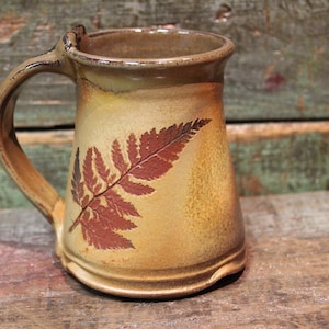 May include: A brown ceramic mug with a fern design. The mug has a handle and a wide base.