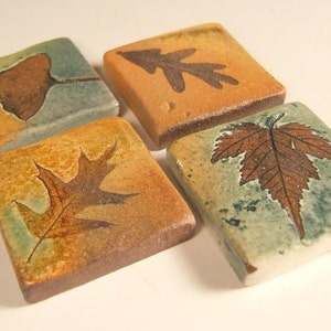 May include: Four ceramic tiles with different leaf designs. Each tile is square and has a light brown background with a darker brown leaf design. The leaves are a ginkgo leaf, an oak leaf, a maple leaf, and a leaf with a more abstract shape.
