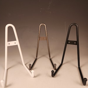 May include: Three metal plate stands in white, silver, and black. Each stand has a curved, A-shaped design with rounded ends.