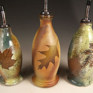 Puede incluir: Tres botellas de aceite de oliva de cerámica con diferentes diseños de hojas. Las botellas son marrones, verdes y amarillas con una tapa de plata metálica.