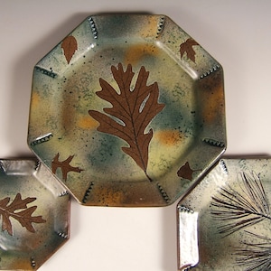 Op de afbeelding: Drie keramische platen met een achthoekige vorm. De platen zijn versierd met een groene, bruine en witte glazuur. De middelste plaat heeft een groot bruin blad, terwijl de kleinere platen een kleiner bruin blad en een bruine dennennaald hebben.