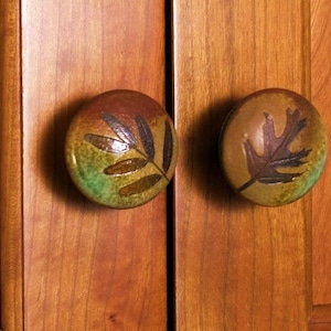 May include: Two ceramic cabinet knobs with leaf designs. The knob on the left has a green, brown, and gold leaf design. The knob on the right has a brown and gold leaf design.