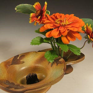 Może przedstawiać: Ceramiczny wazon z brązową i zieloną glazurą, ozdobiony wzorem liści. Wazon jest wypełniony pomarańczowymi i żółtymi kwiatami.