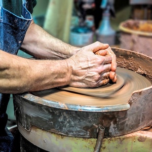 May include: A potter's hands shaping a lump of clay on a spinning pottery wheel. The clay is a reddish-brown color and the wheel is made of metal.