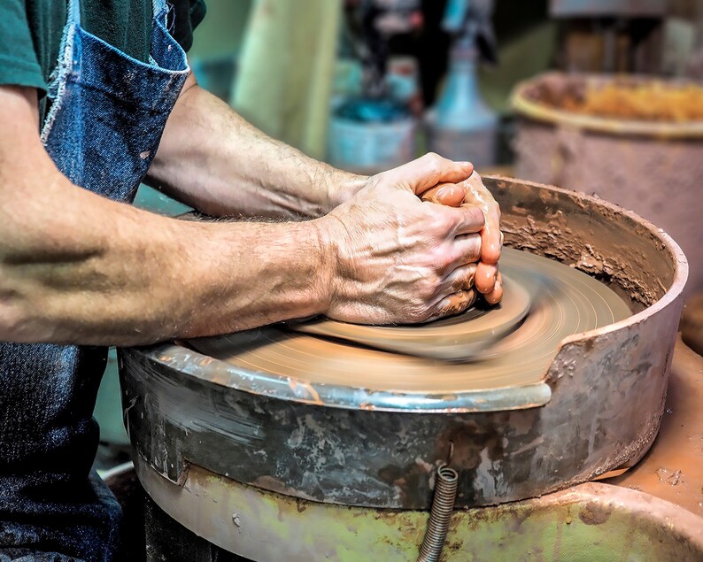 May include: A potter's hands shaping a lump of clay on a spinning pottery wheel. The clay is a reddish-brown color and the wheel is made of metal.