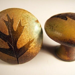 May include: Two ceramic knobs with a leaf design. The knob on the left has a dark brown leaf on a light brown, green, and blue background. The knob on the right has a dark brown leaf on a light brown background.