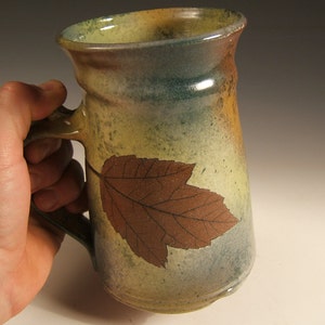 Può includere: Una tazza in ceramica con un design a foglia marrone su uno sfondo verde e giallo. La tazza ha un manico ed è realizzata in stile rustico.