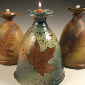 May include: Three ceramic oil lamps with leaf designs. The lamps are glazed in shades of brown, green, and tan. The centre lamp has a maple leaf design, the left lamp has a fern design, and the right lamp has an oak leaf design.