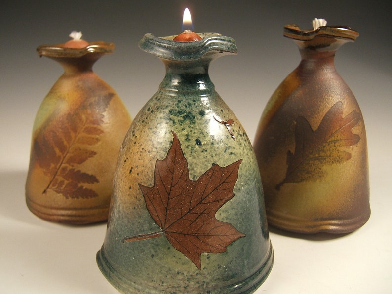 May include: Three ceramic oil lamps with leaf designs. The lamps are glazed in shades of brown, green, and tan. The centre lamp has a maple leaf design, the left lamp has a fern design, and the right lamp has an oak leaf design.