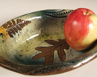 May include: Handmade ceramic serving bowl with a leaf design. The oval bowl has a speckled green and brown glaze. A red and yellow apple rests inside the bowl. The bowl has a rustic, natural aesthetic.