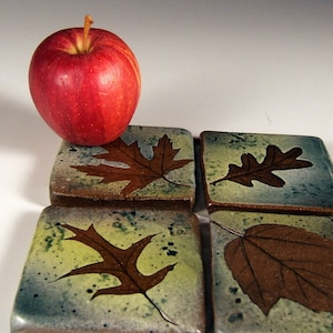 Puede incluir: Cuatro posavasos cuadrados de cerámica con diseños de hojas marrones sobre un fondo azul y verde. Una manzana roja se encuentra encima de los posavasos.