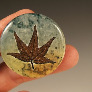 Könnte beinhalten: Eine runde Keramiknadel mit einem braunen Ahornblatt-Design auf blauem und gelbem Hintergrund.